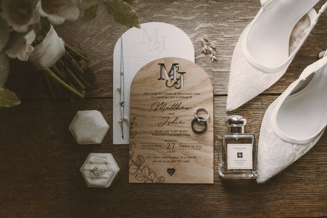 A styled flat-lay of wedding stationery and accessories on a rustic wooden surface, including an arch card, a wooden invitation with monogram lettering, two rings, a small perfume bottle, and white lace wedding shoes.