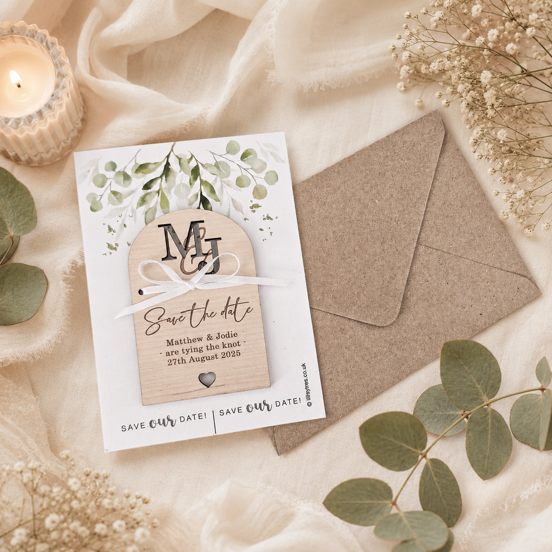 A styled flat-lay of a white 'Save the Date' card with botanical leaf illustrations and a wooden monogram tab, placed beside a kraft paper envelope on cream fabric with dried flowers and greenery.